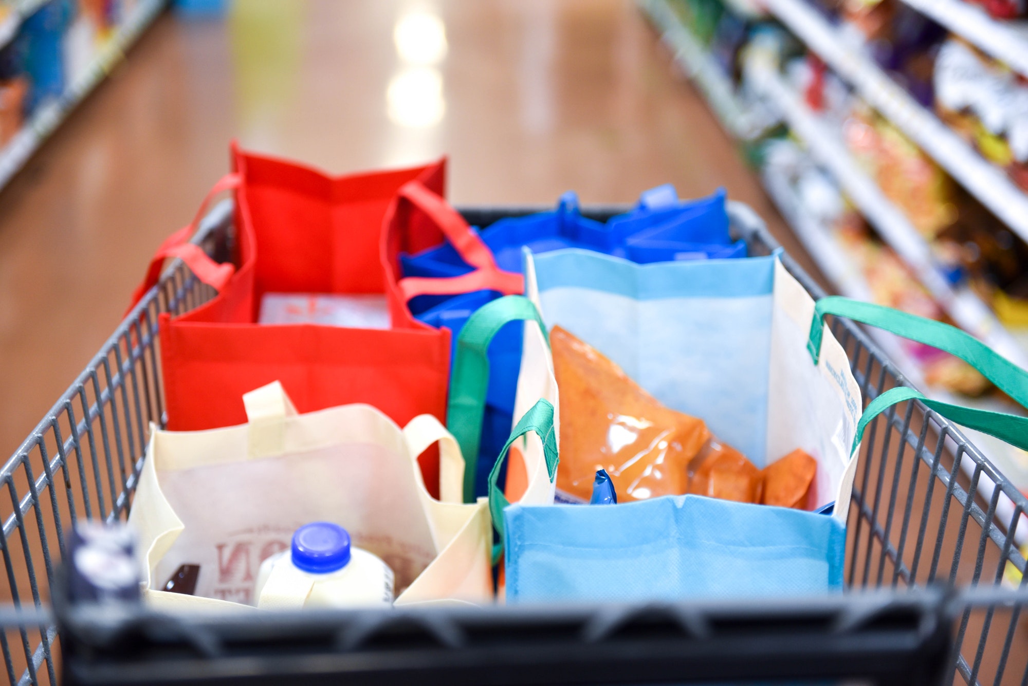 Close-up image of a shopping cart in the middle of a grocery store isle filled with reusable